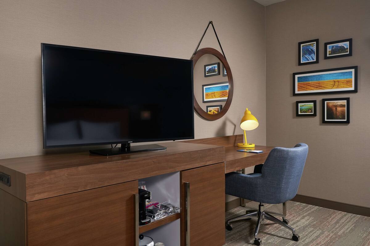 A hotel room with a large flat-screen TV, a wooden desk, a blue office chair, a desk lamp, and several framed pictures on the wall.