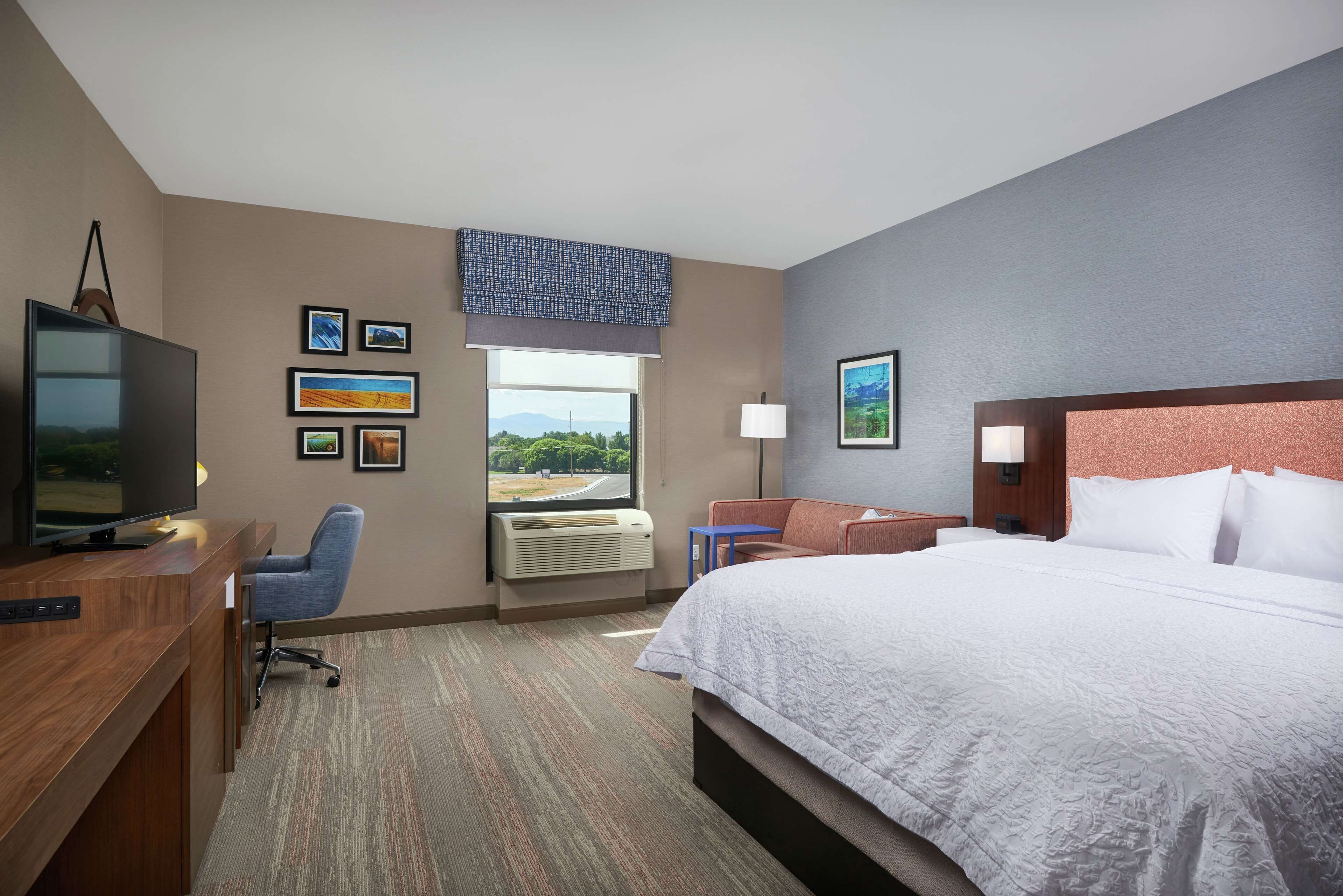 A hotel room with a large bed, a desk with a TV, a chair, a window with a view, and several framed pictures on the wall.