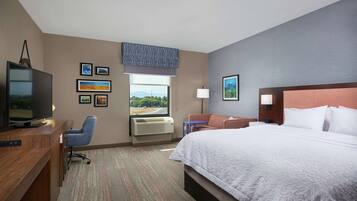 A hotel room with a large bed, a desk with a TV, a chair, a window with a view, and several framed pictures on the wall.