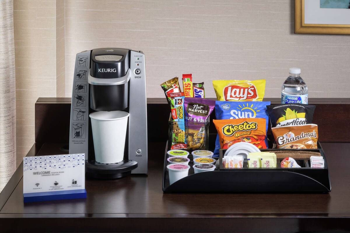 A coffee maker, a tray of snacks, and a welcome sign on a hotel counter.