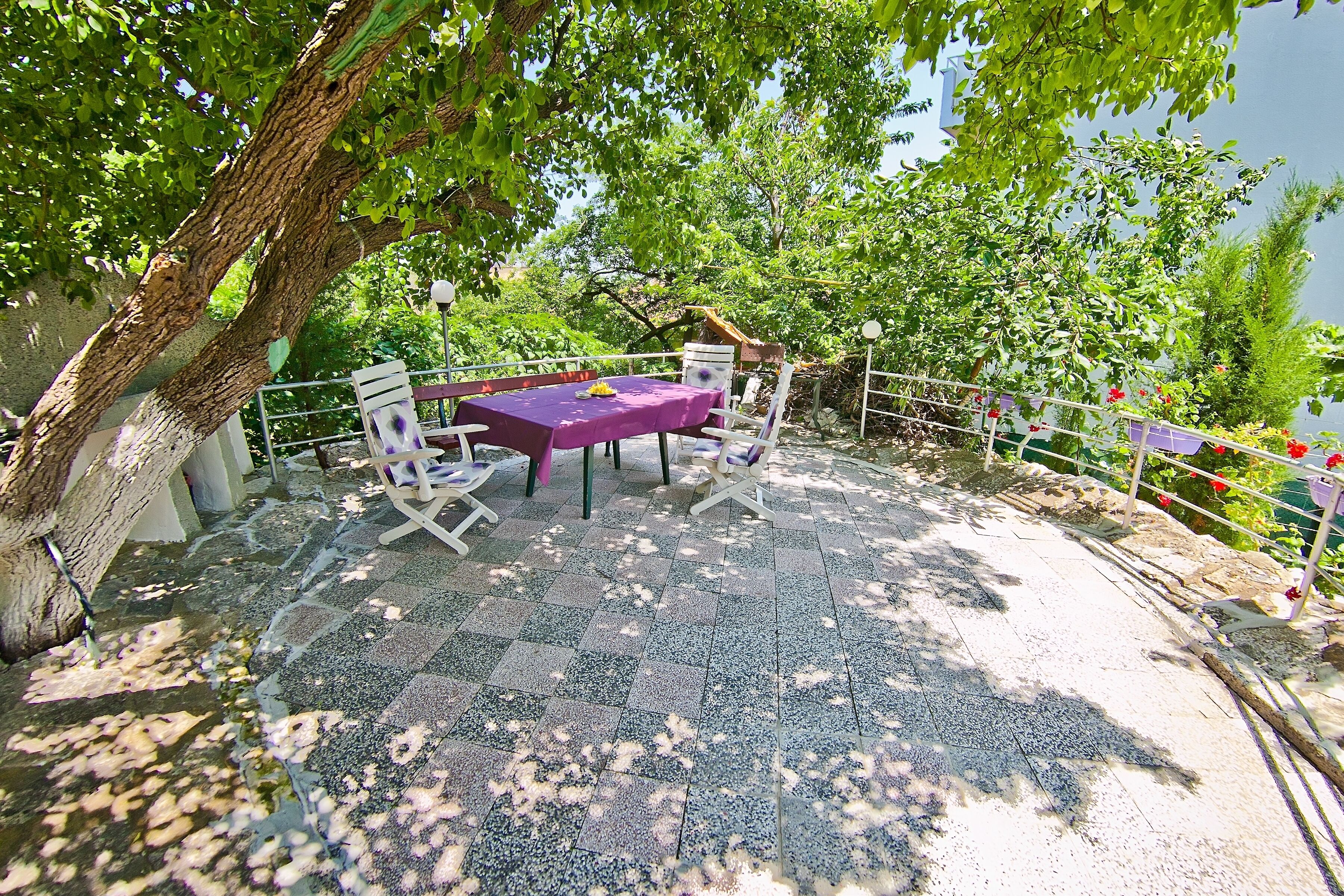 An outdoor seating area with a table and chairs under a tree, surrounded by a railing and greenery.
