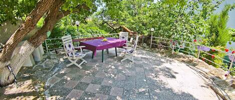 An outdoor seating area with a table and chairs under a tree, surrounded by a railing and greenery.