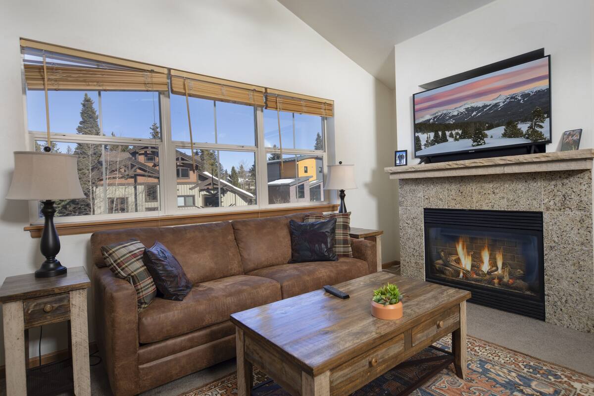 A living room with a sofa, coffee table, fireplace, TV, and large window with blinds.