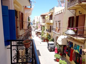 A narrow street with buildings on both sides, balconies with potted plants, and a truck parked on the side.