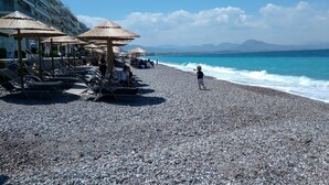 A pebbled beach with umbrellas and lounge chairs, a building with balconies, and the sea in the background. - Luxury apart. w/ spectacular views (Loutraki-Korinthias)