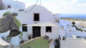 Un bâtiment blanc avec une porte sombre et une petite fenêtre, entouré de plantes en pot et surmonté d’un auvent blanc.