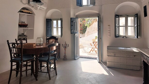 Un coin repas avec une table et des chaises en bois, un lustre et une vue sur l’extérieur grâce à de grandes fenêtres ouvertes.