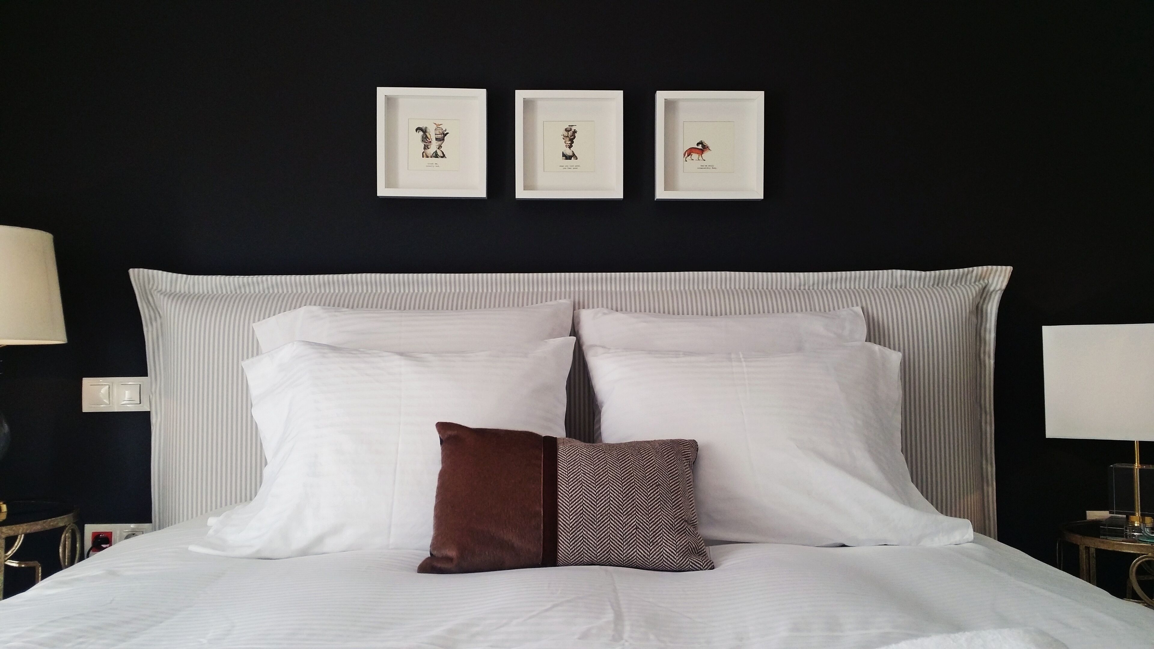 A bed with white linens and a brown pillow, flanked by two bedside tables with lamps, against a dark wall with three framed pictures.