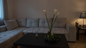 Olohuoneessa on kulmasohva, sohvapöydällä on maljakko täynnä kukkia ja sivupöydällä on lamppu.