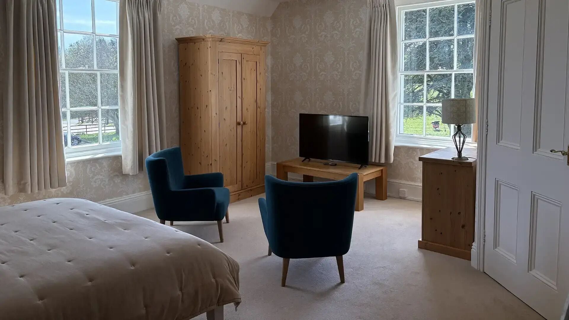 Un dormitorio con una cama, dos sillones azules, un armario de madera, un televisor sobre una mesa de madera y una lámpara.