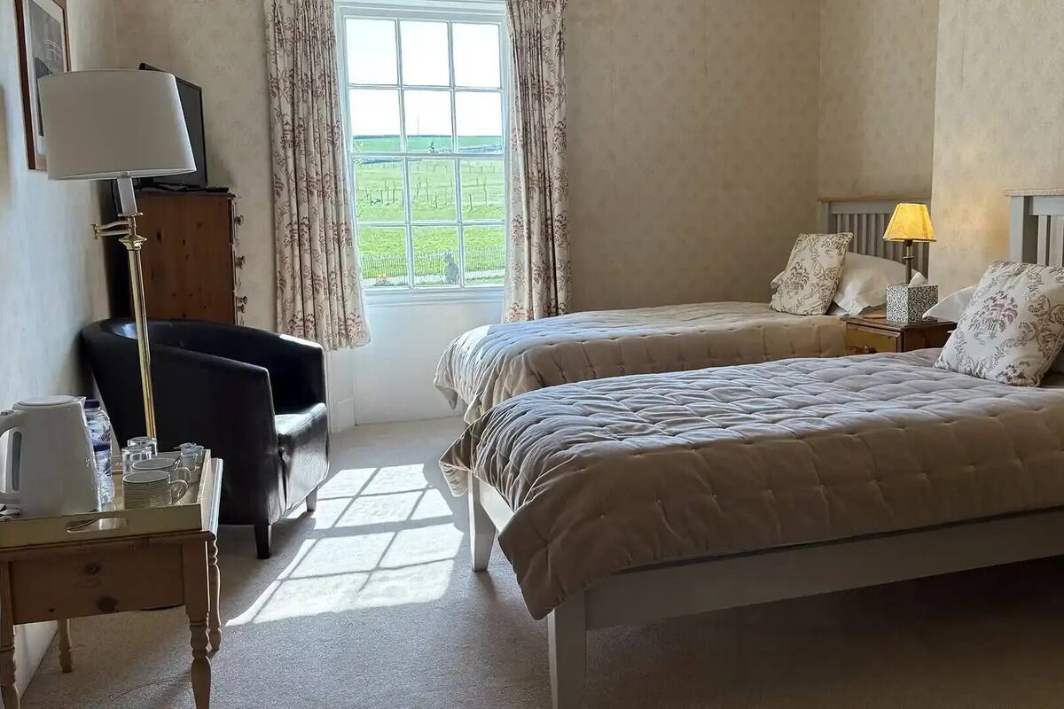 Un dormitorio con dos camas, un escritorio con lámpara, una silla y una ventana con cortinas.