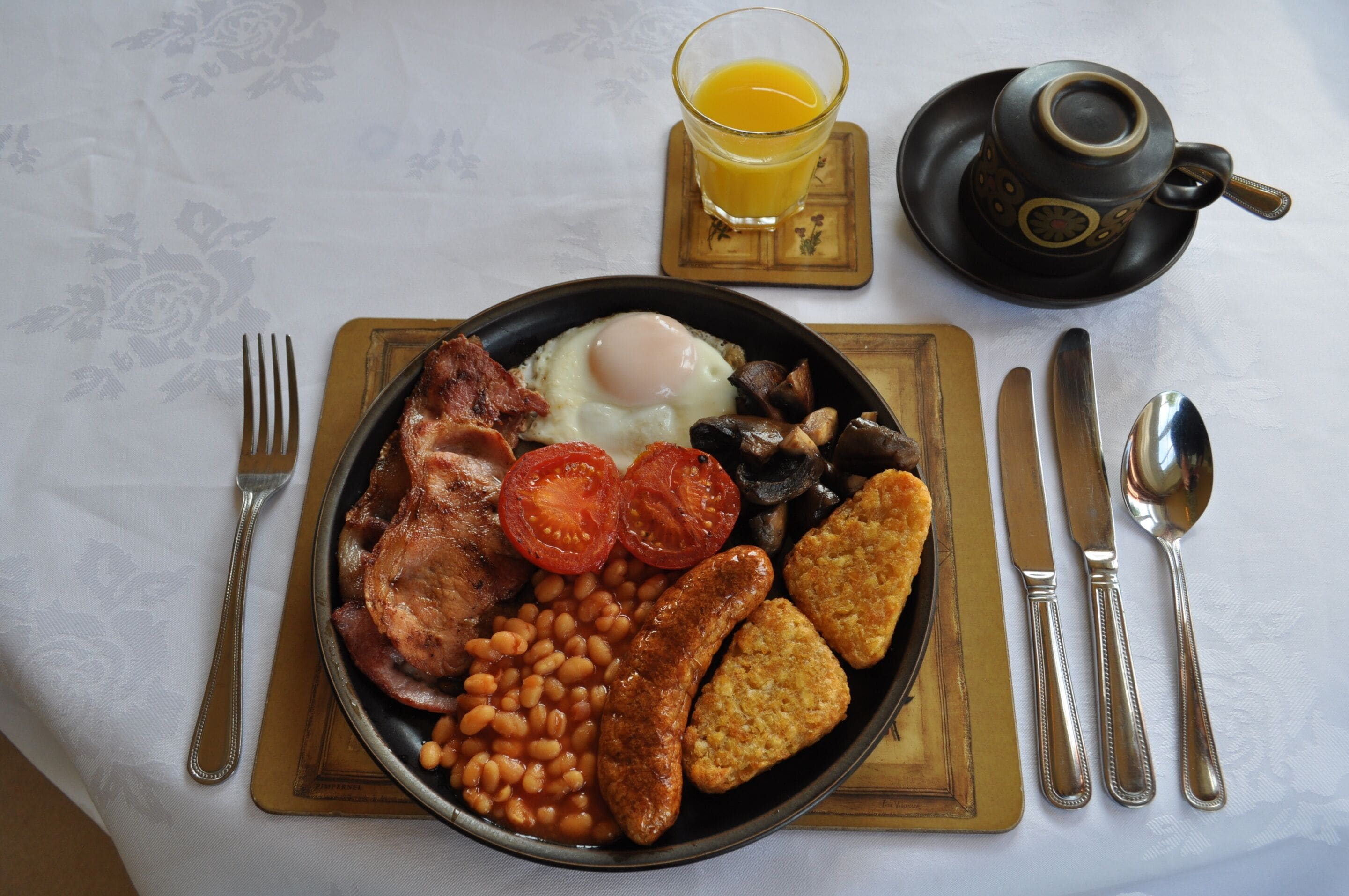A breakfast plate with eggs, bacon, tomatoes, beans, sausages, and toast, accompanied by a glass of orange juice and a cup of tea on a table with a floral tablecloth.