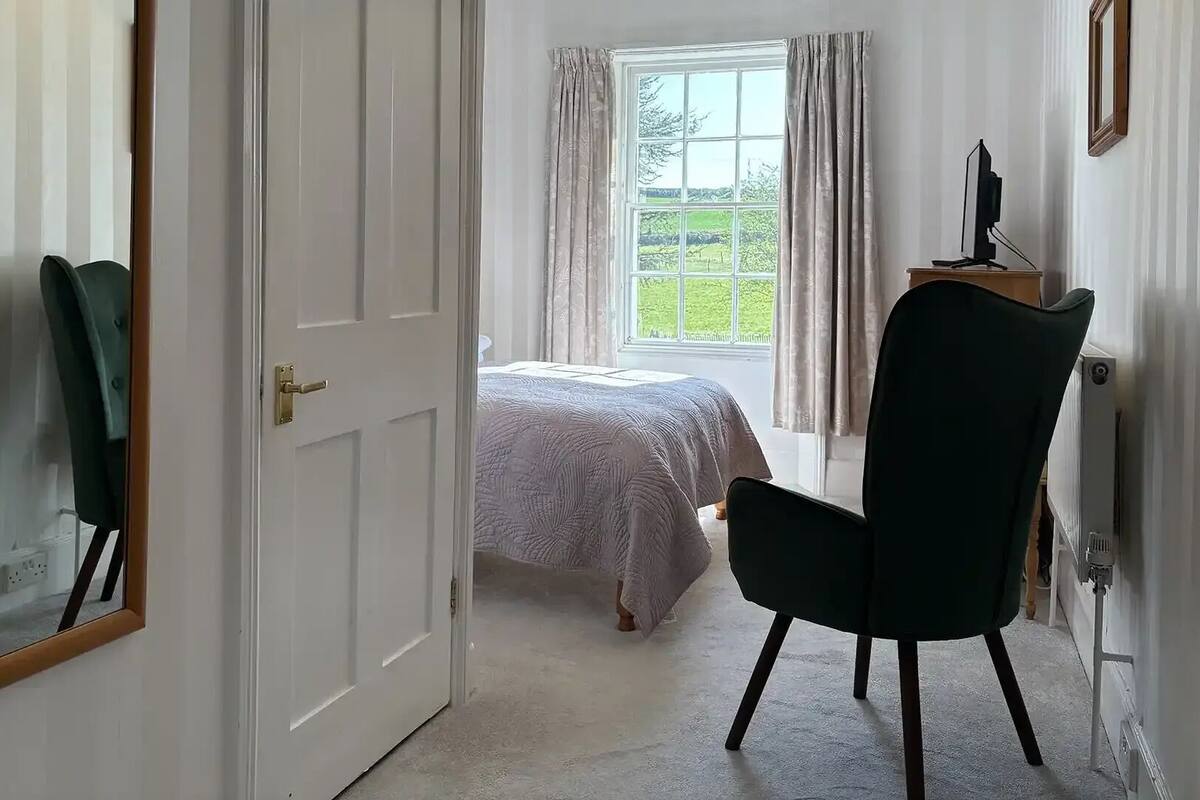 Un dormitorio con cama, una silla, un escritorio y una ventana con cortinas.