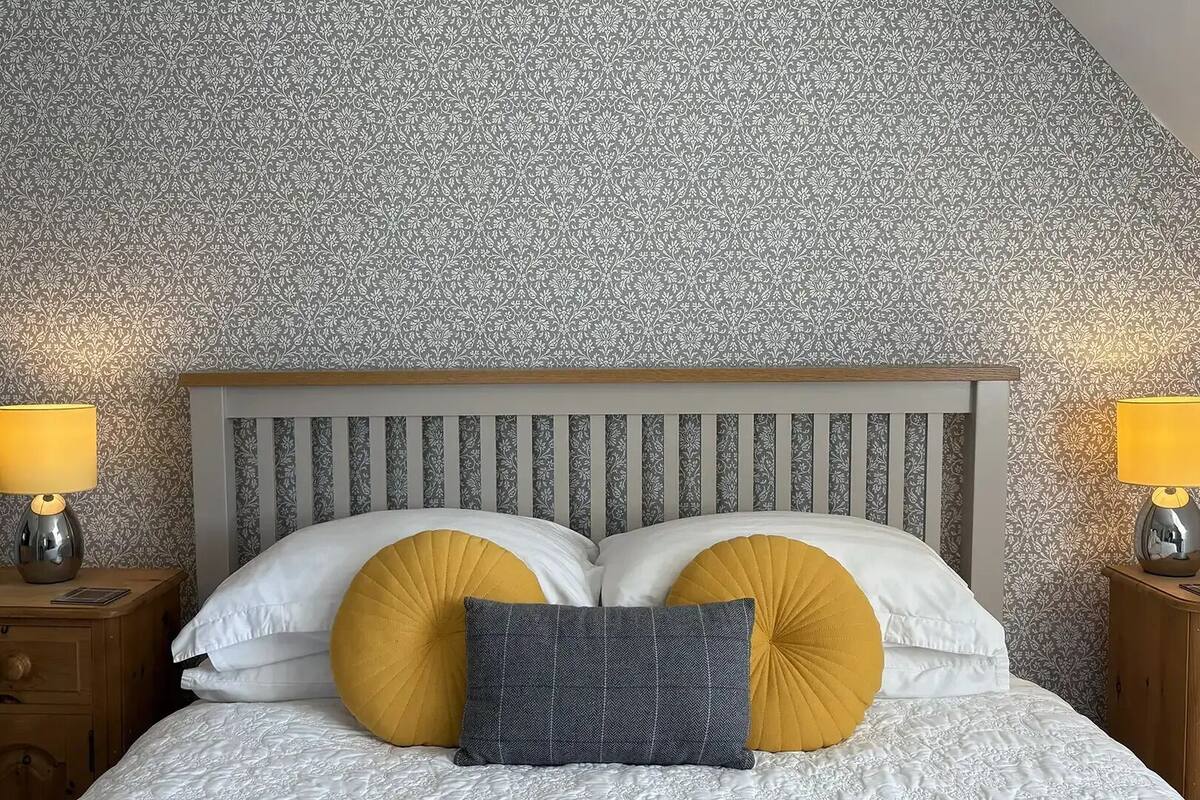 Un dormitorio con papel tapiz estampado, cabecera de madera, dos mesitas de noche con lámparas y una cama con almohadas blancas y mostaza.