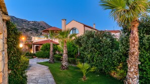 A stone pathway leading to a house with a red roof, surrounded by lush greenery and palm trees. - Luxury stone villa with outdoor jacuzzi, private pool and antique furniture (Πλάκα)