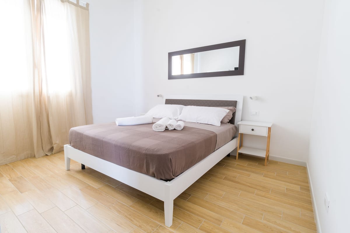 Cama bem arrumada com roupa de cama branca e castanha, mesa de cabeceira e espelho na parede.
