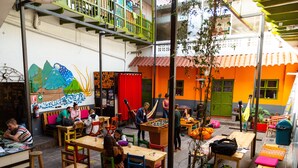 Un café al aire libre con mesas y sillas, un mural en la pared y un edificio de paredes anaranjadas y verdes al fondo.