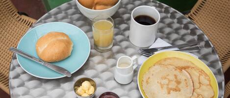 A breakfast table with a plate of pancakes, a bowl of croissants, a glass of orange juice, a cup of coffee, and a small bowl of butter.