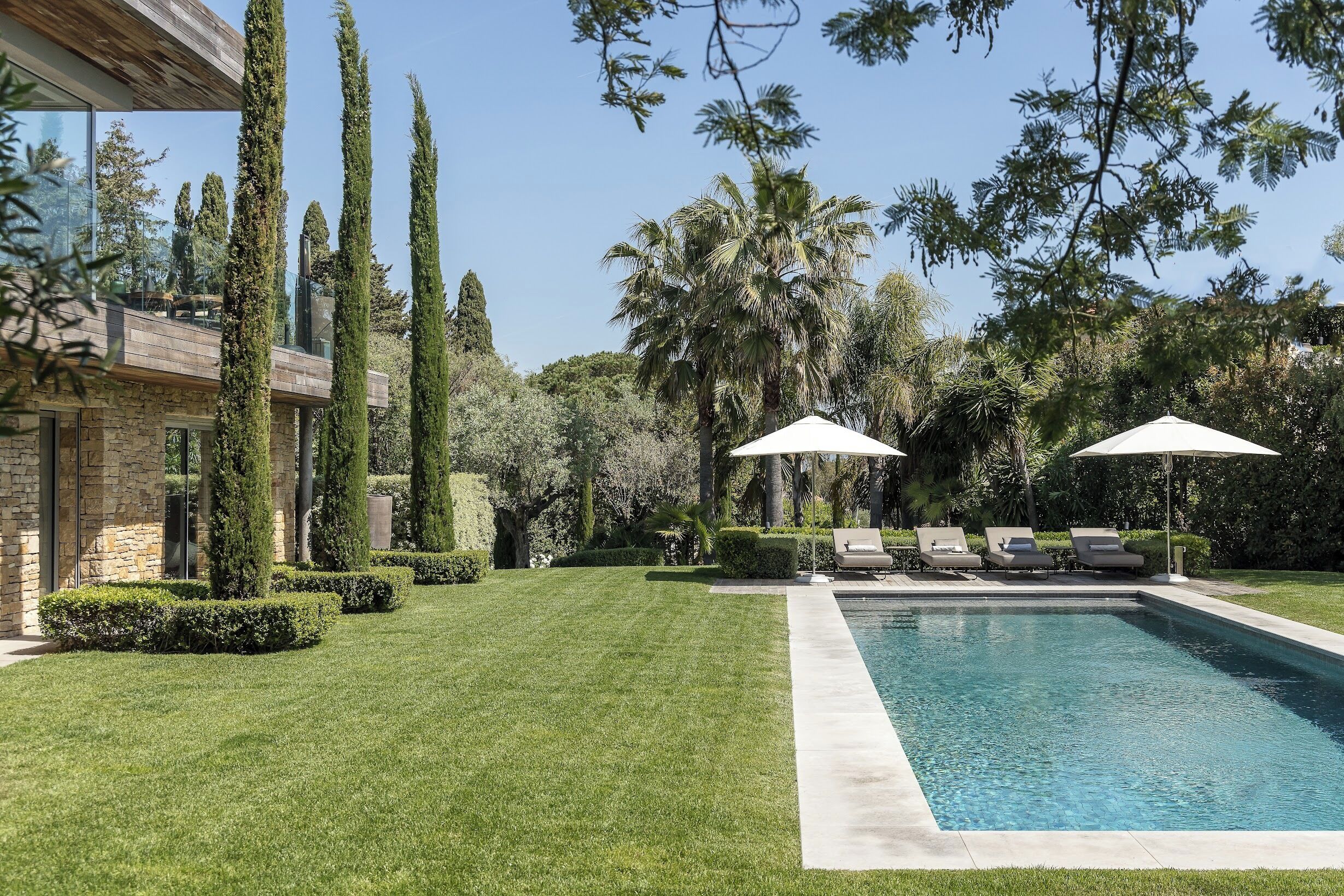 A modern pool area with a clear blue pool, surrounded by well-manicured grass and tall trees. There are lounge chairs and umbrellas under the trees.