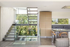 A modern interior with a glass staircase leading to a balcony, a kitchen area with bar stools, and a view of greenery.