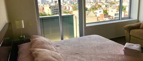 Habitación de hotel con una cama grande, una mesita de noche con lámpara, una silla y vistas a un paisaje urbano a través de amplios ventanales.