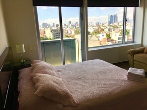 A hotel room with a large bed, a nightstand with a lamp, a chair, and a view of a cityscape through large windows. - Napoles Condo Suite (Mexico City)