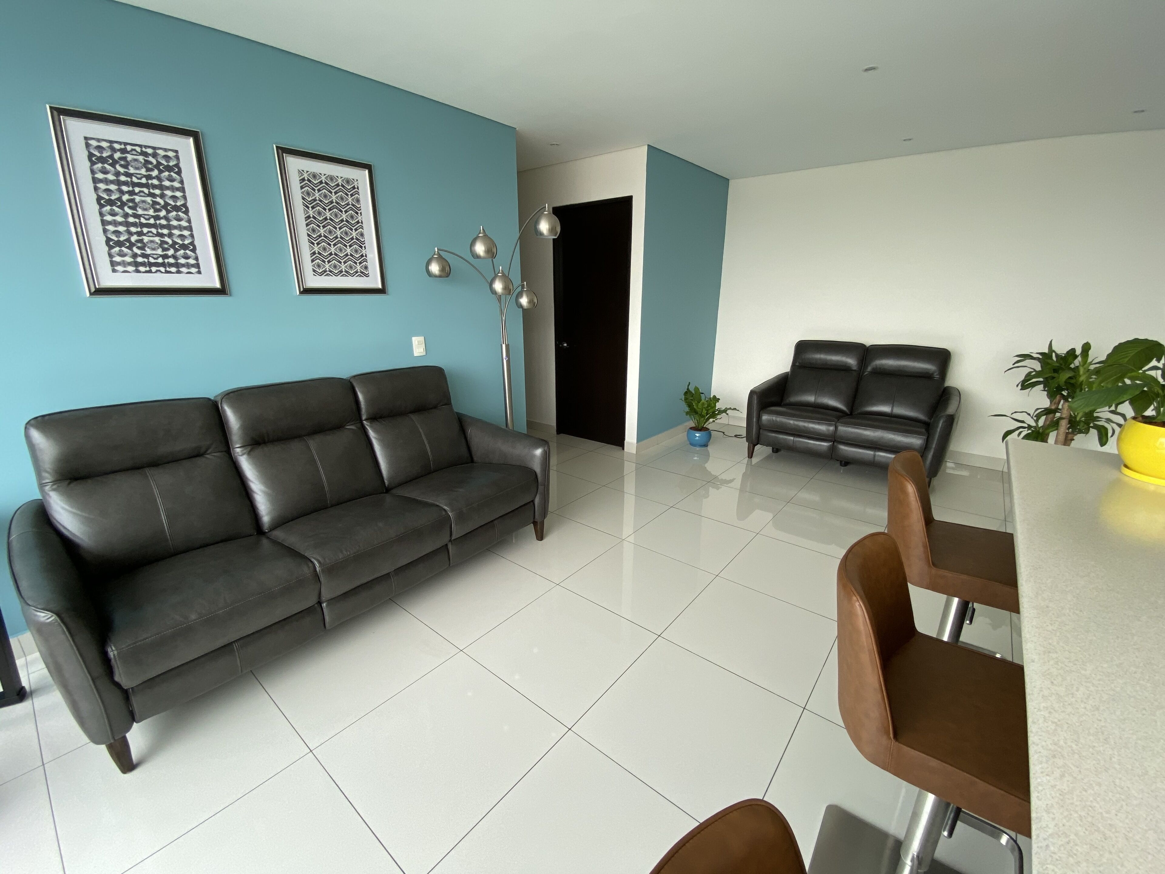 Una sala de estar moderna con un sofá de cuero negro, una mesa de comedor con sillas y dos cuadros en la pared.