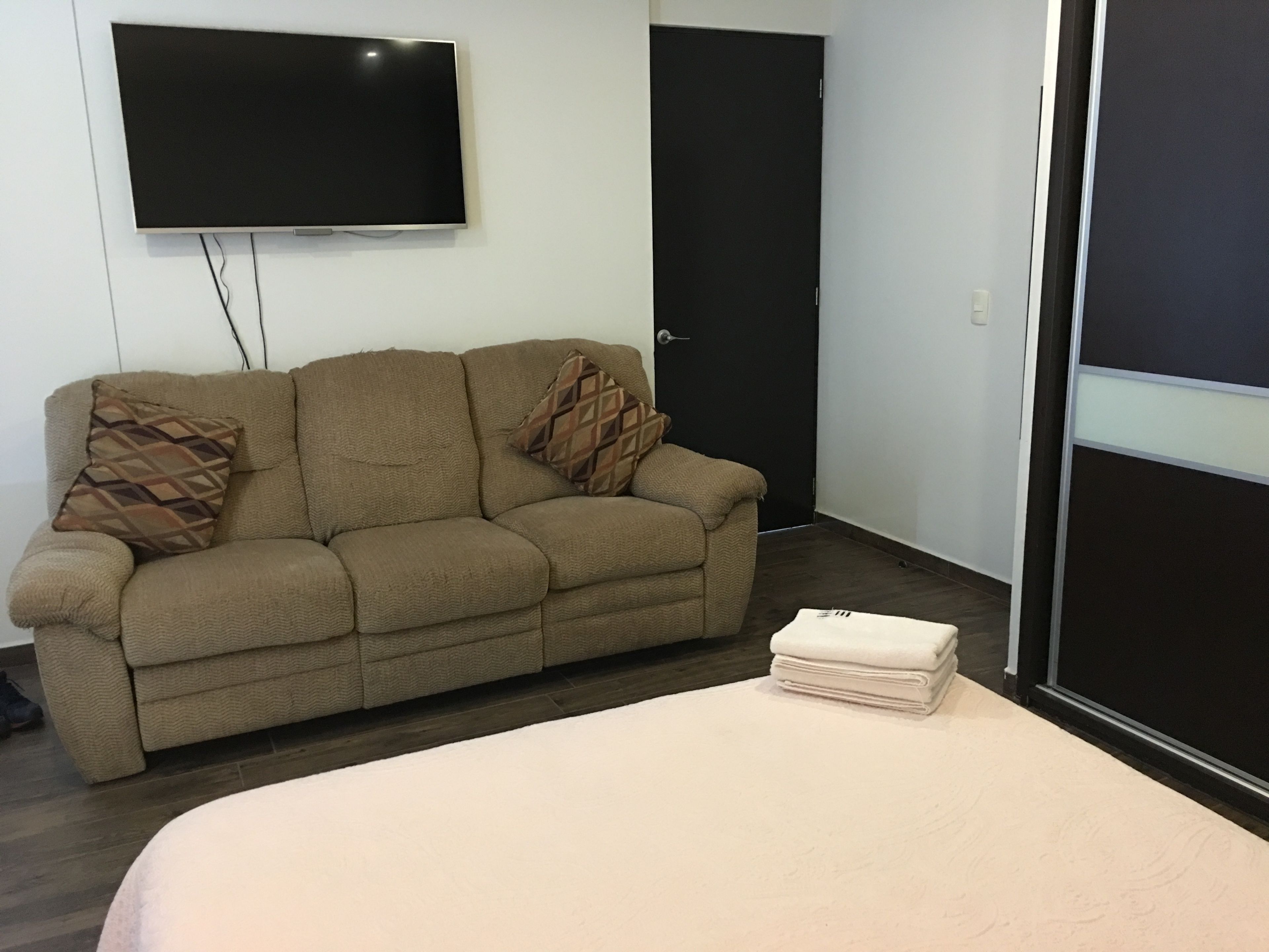 Een kamer met een beige bank, een flatscreen televisie aan de muur, een zwarte deur en een bed met een witte deken.