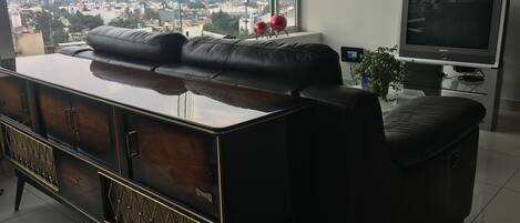 Una sala moderna con un sofá de cuero negro grande, un mueble de entretenimiento de madera y un televisor sobre un pedestal.
