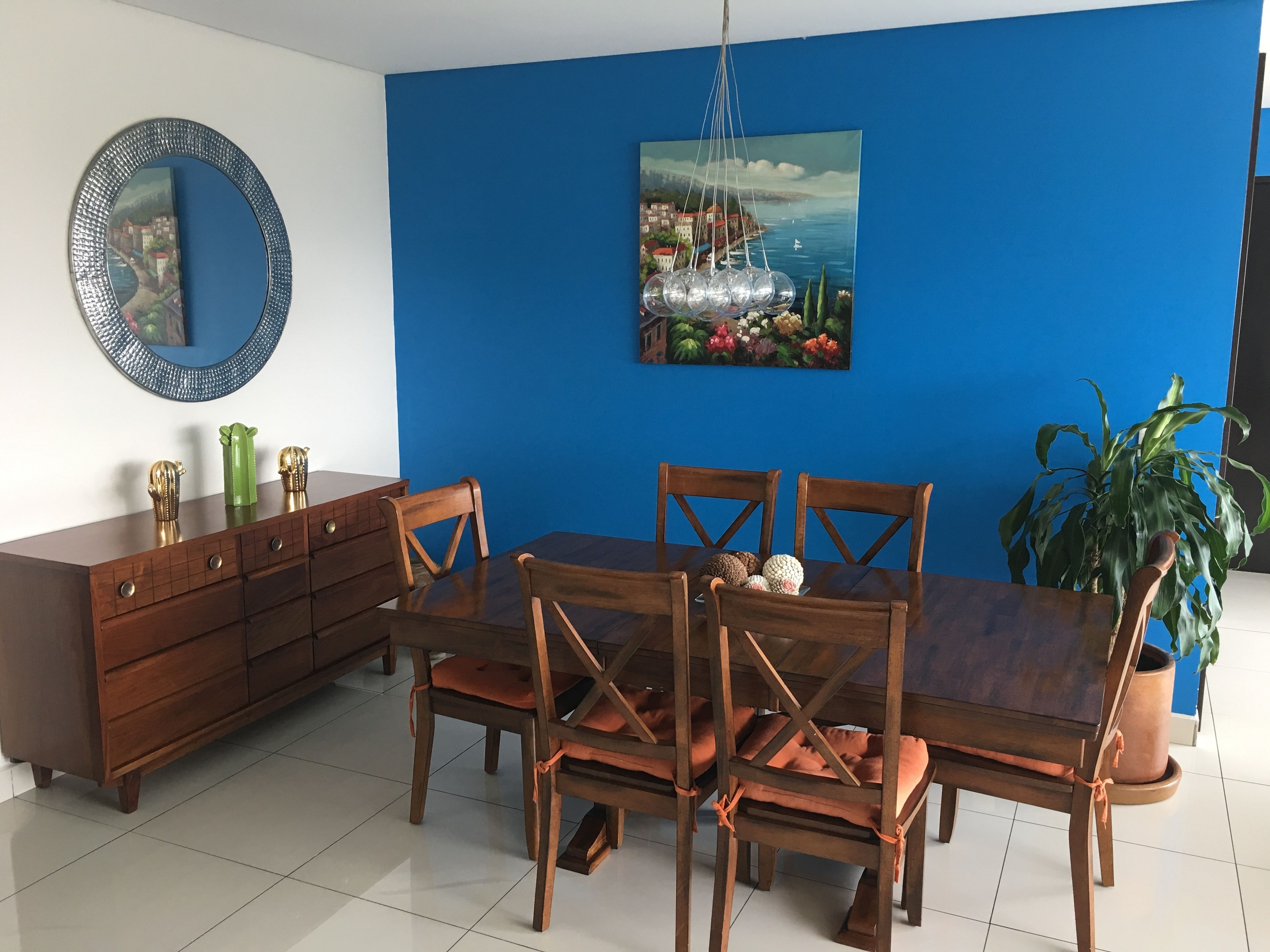 A dining area with a wooden table and chairs, a blue accent wall, a decorative mirror, and a potted plant.