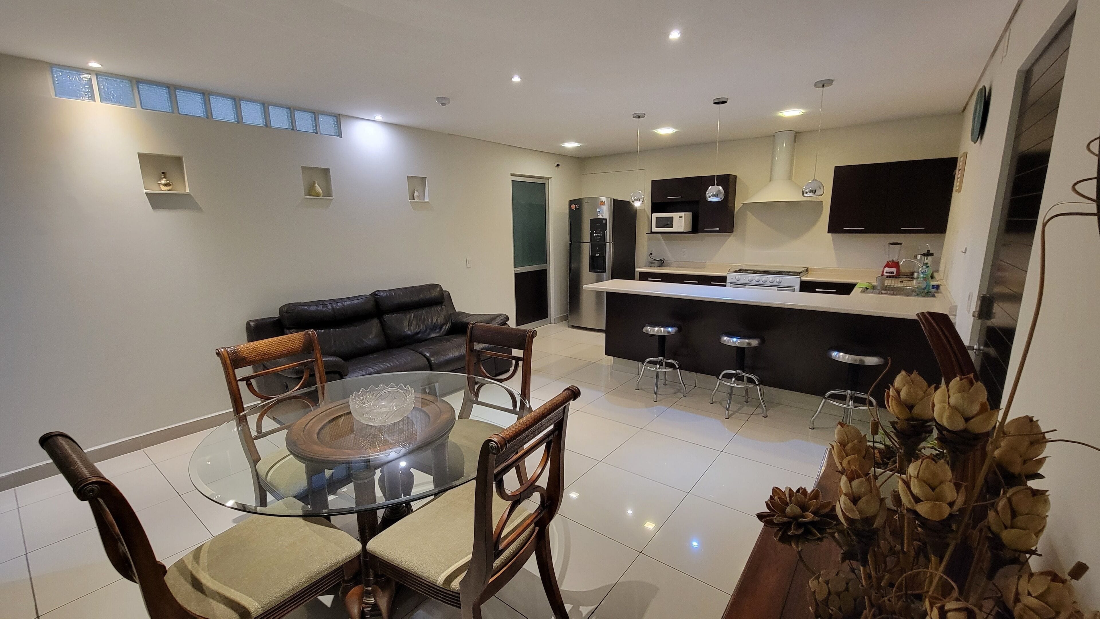 A modern kitchen with a central island, a dining area with a glass table and chairs, and a living room with a black leather sofa.