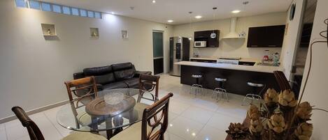 A modern kitchen with a central island, a dining area with a glass table and chairs, and a living room with a black leather sofa.