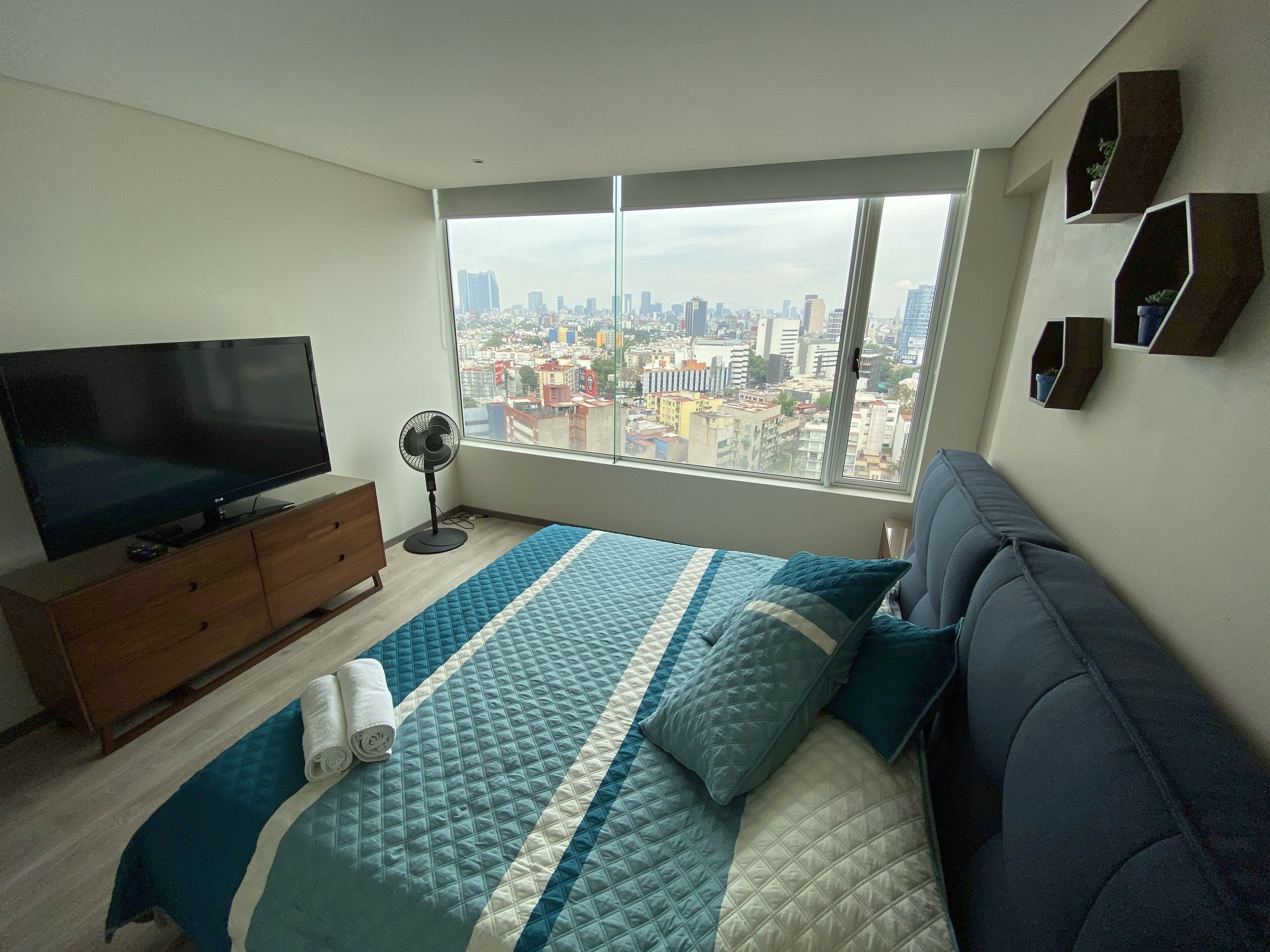 Een slaapkamer met een groot raam, een bed, een televisie, een ventilator en een dressoir.