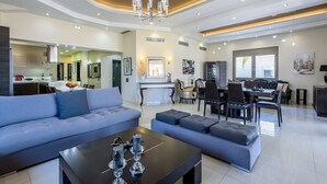 Ein großzügiger Wohnbereich mit einem modernen Sofa, einem Hocker und einem Couchtisch. Es gibt einen Essbereich mit Tisch und Stühlen, und im Hintergrund befindet sich eine Küche.