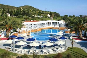 A resort pool area with sun loungers and umbrellas, surrounded by palm trees and a hillside in the background. - Ioli Village (Kassandra)