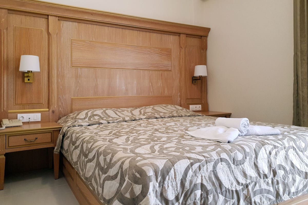 A hotel room with a wooden headboard, two bedside lamps, a telephone, and a neatly made bed with a patterned bedspread.