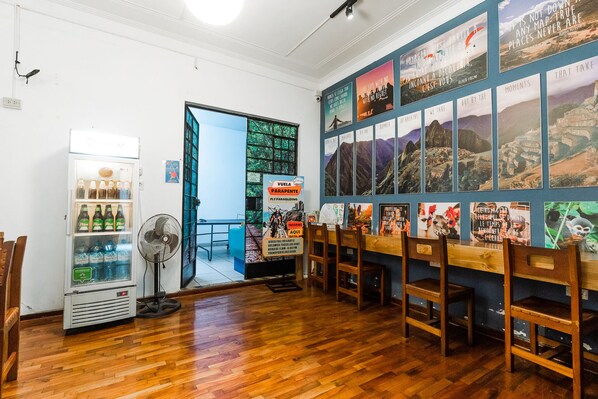 Interior de restaurante con sillas de madera, un ventilador, un refrigerador con bebidas y una pared con carteles de viajes.