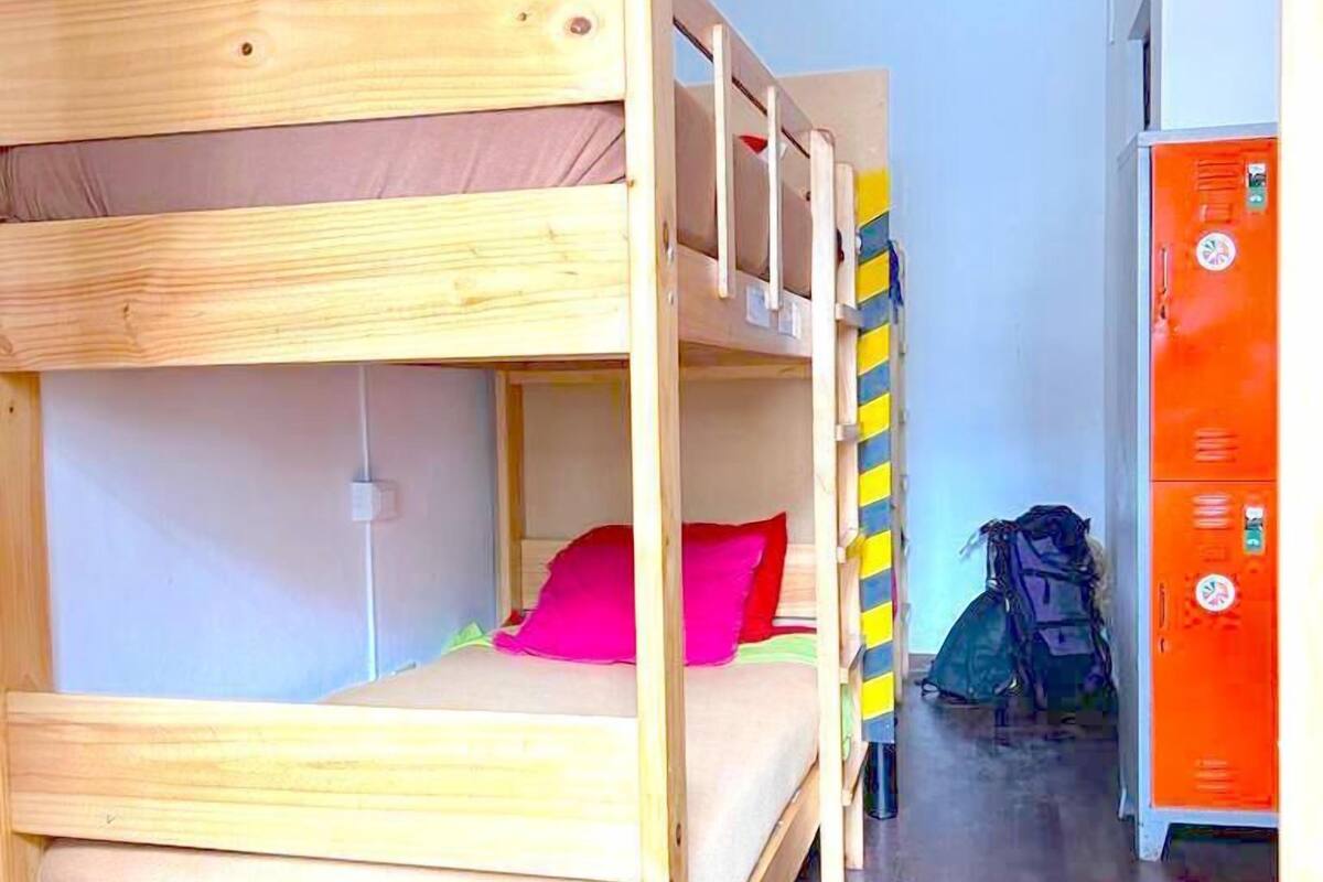 Un lit superposé avec une structure en bois, un petit bureau et une chaise. Il y a une armoire avec un miroir et un porte-manteau.