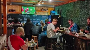 A casual dining area with wooden tables and stools, a chalkboard menu, and a television displaying a sports game. - Dragonfly Hostels Miraflores (Lima)