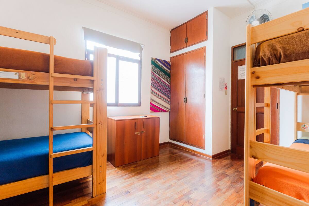 Une chambre de dortoir avec des lits superposés, une armoire en bois, une fenêtre et une tapisserie aux couleurs vives.