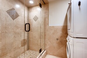 A modern bathroom with a glass shower enclosure, a built-in washing machine, and decorative mosaic tiles on the walls.