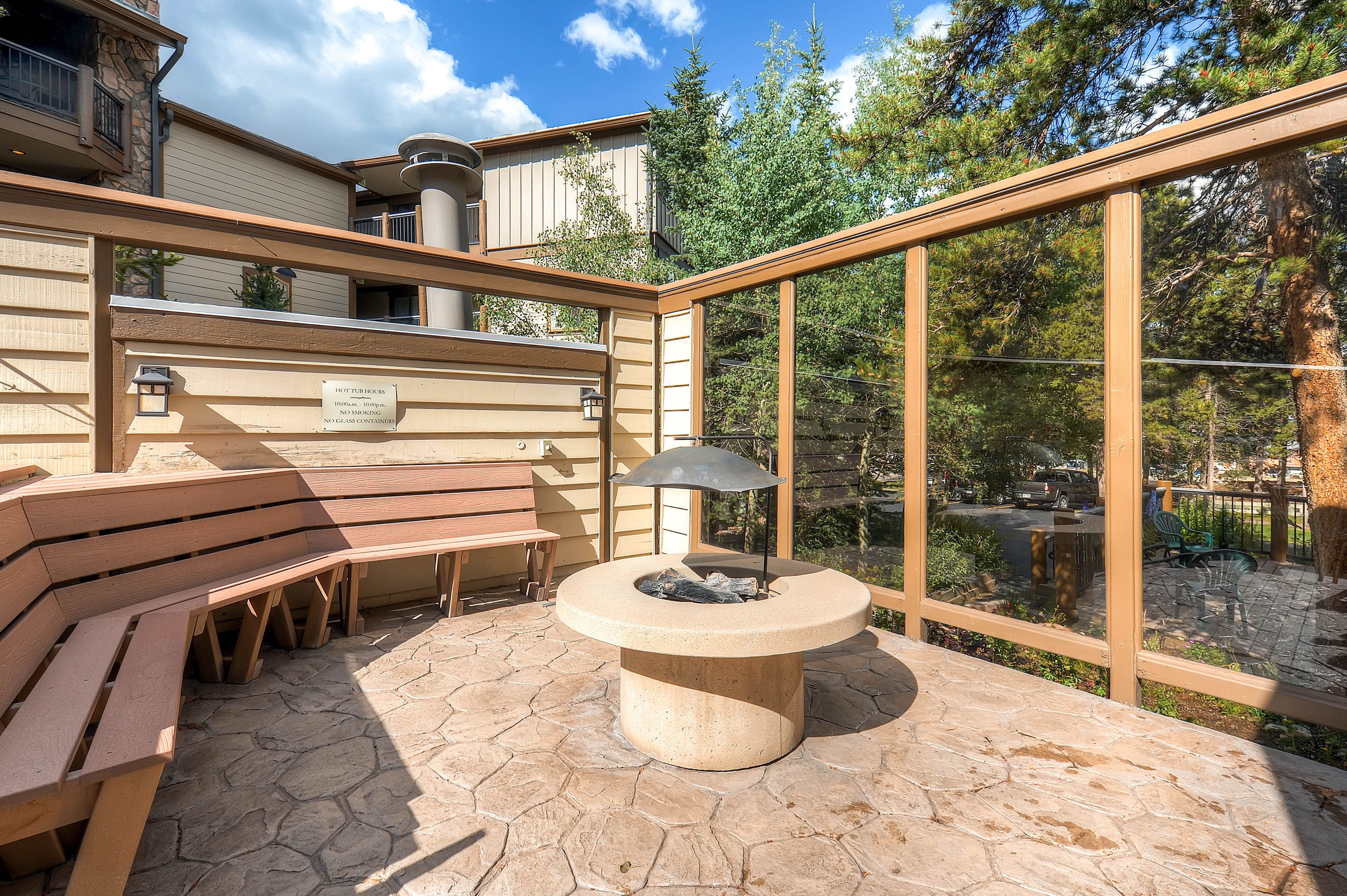 Eine Terrasse mit Holzbänken und einem Steinkamin, umgeben von einer Glasveranda mit Blick auf Bäume und ein Gebäude.