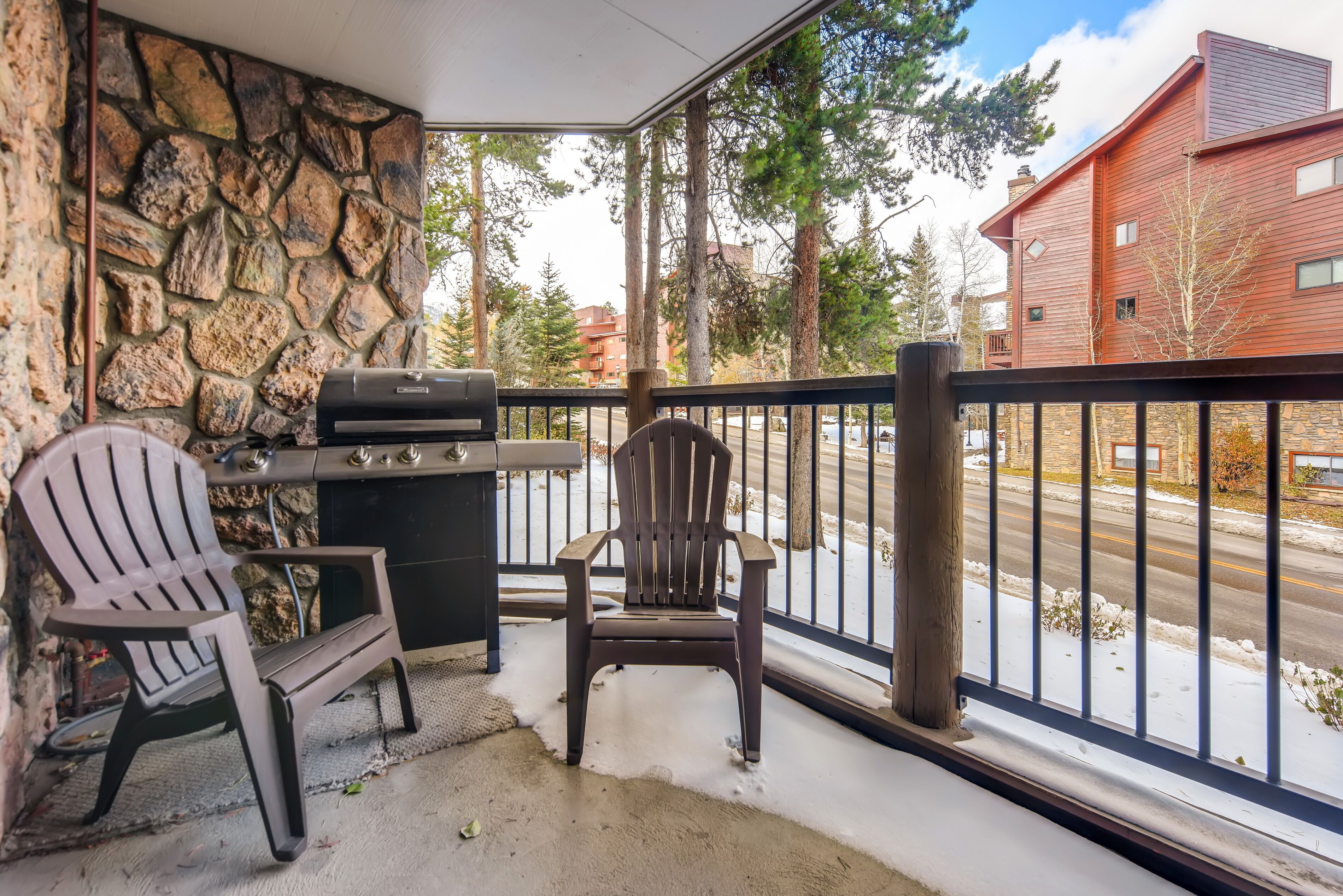 Ein Balkon mit Steinwänden, einem Grill, zwei Adirondack-Stühlen und Blick auf eine verschneite Straße und Gebäude.