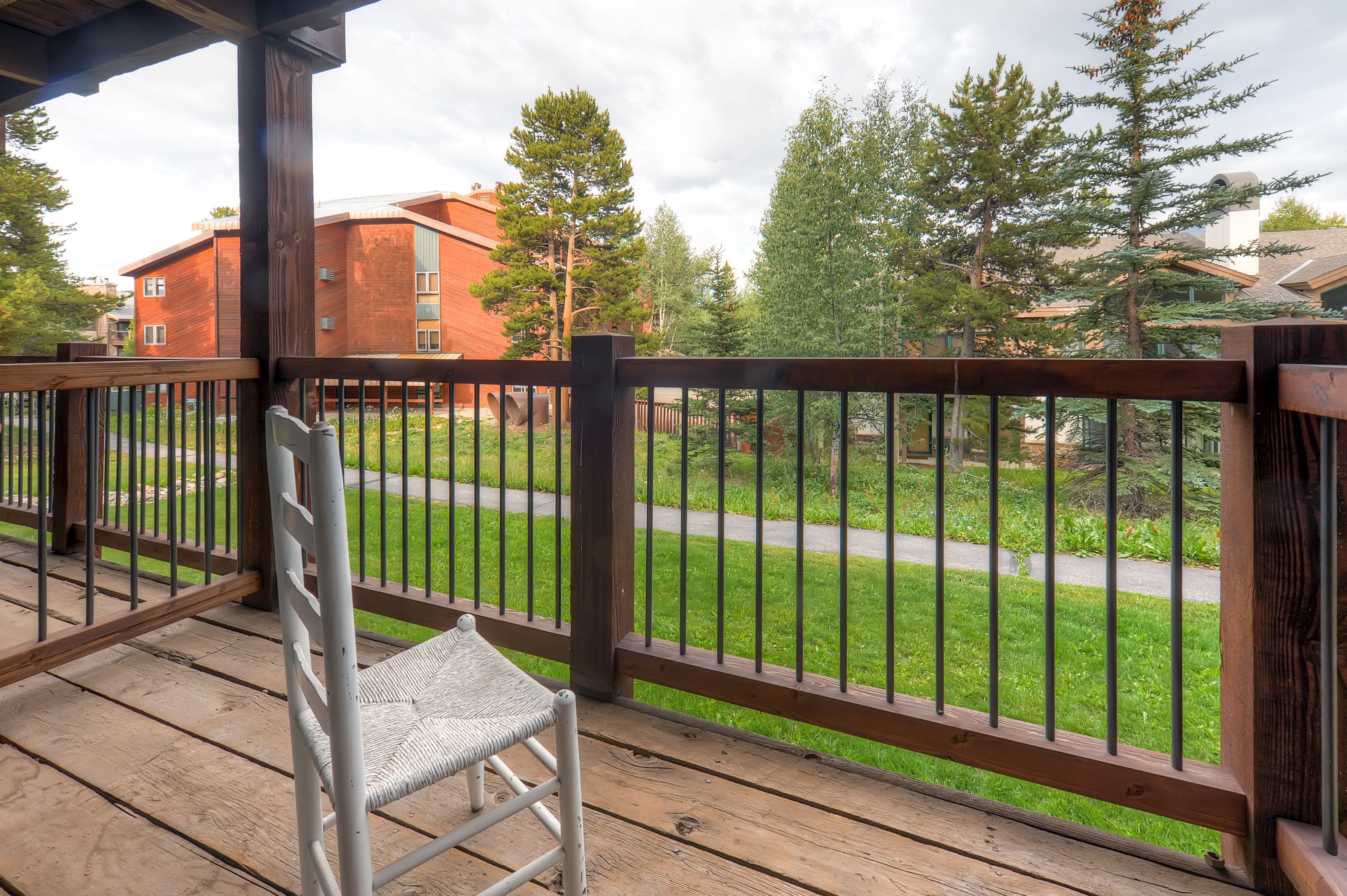 A wooden deck with a metal railing, a white chair, and a view of a green lawn and buildings in the background.