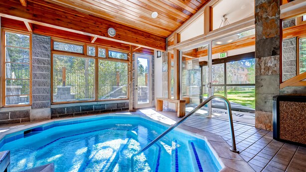Une piscine intérieure à l’eau d’un bleu cristallin, avec des poutres en bois au plafond, des murs en pierre et de grandes fenêtres offrant une vue sur l’extérieur.