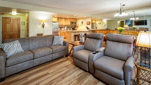 Un salon avec un canapé, un fauteuil inclinable et une table basse. Au fond, on aperçoit une cuisine avec des meubles en bois et un réfrigérateur.