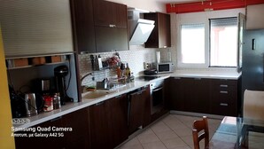Una cucina con pensili in legno scuro, un frigorifero in acciaio inossidabile, un forno a microonde e un lavello. Si vede una finestra con persiane e un tavolo con sedie.