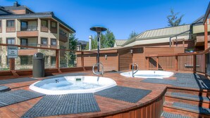 A rooftop spa area with two hot tubs, wooden deck, and a view of surrounding buildings.