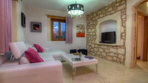 A living room with a stone wall, a white sofa, a coffee table with a laptop, and a TV mounted on the wall. - Green Paradise Luxury Villa In Crete  (Τζαννακιανά)