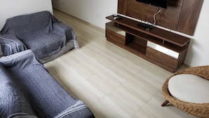 A living room with a sofa, a TV stand with a flat-screen TV, a wicker chair, and a small round table. - Apartamento Central Setti (Foz do Iguaçu)